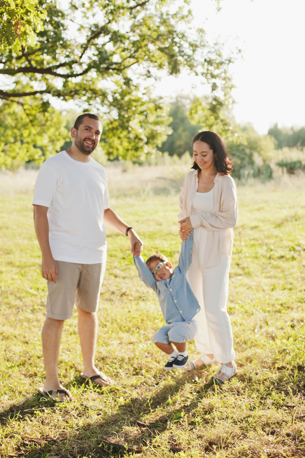 A smiling man and woman stand in a sunny grassy area, each holding onto a child who is suspended between them, joyfully swinging. The man wears a white t-shirt and shorts, while the woman is dressed in light-colored clothing. The child, wearing a denim shirt and sunglasses, is laughing as they are lifted off the ground. In the background, a tree and a blurred landscape can be seen, indicating a bright, outdoor setting.