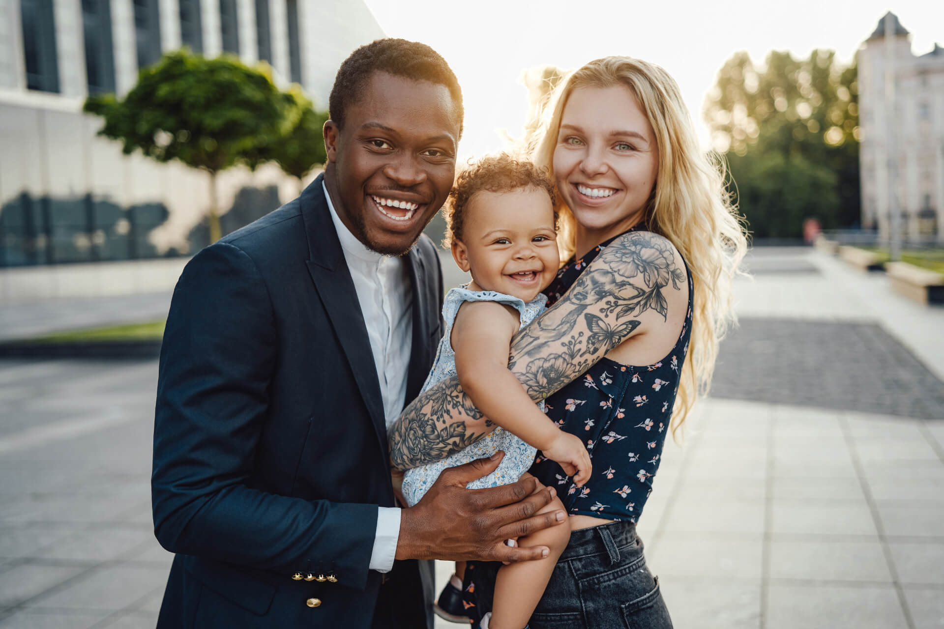 A smiling couple stands together outdoors, holding a joyful baby. The man, wearing a dark suit jacket over a white shirt, and the woman, dressed in a floral top, both have light skin tones and are smiling broadly. The baby, wearing a light blue outfit, is nestled in the woman's arms, grinning happily. The background features modern architecture and greenery, illuminated by soft sunlight.