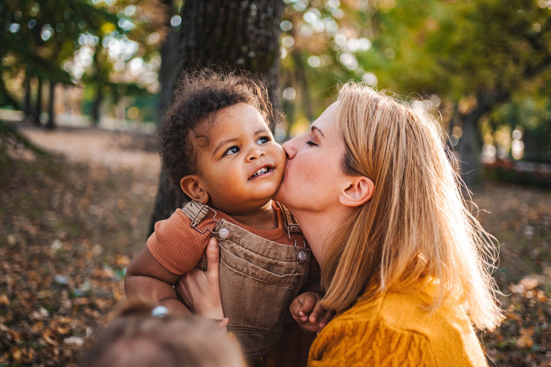 A woman with blonde hair is gently kissing the cheek of a smiling toddler with curly hair, who is being held in her arms. They are surrounded by a scenic outdoor setting with trees and fallen leaves in the background, creating a warm and joyful atmosphere. The woman is wearing a cozy sweater, and the child is dressed in a casual outfit with overalls.