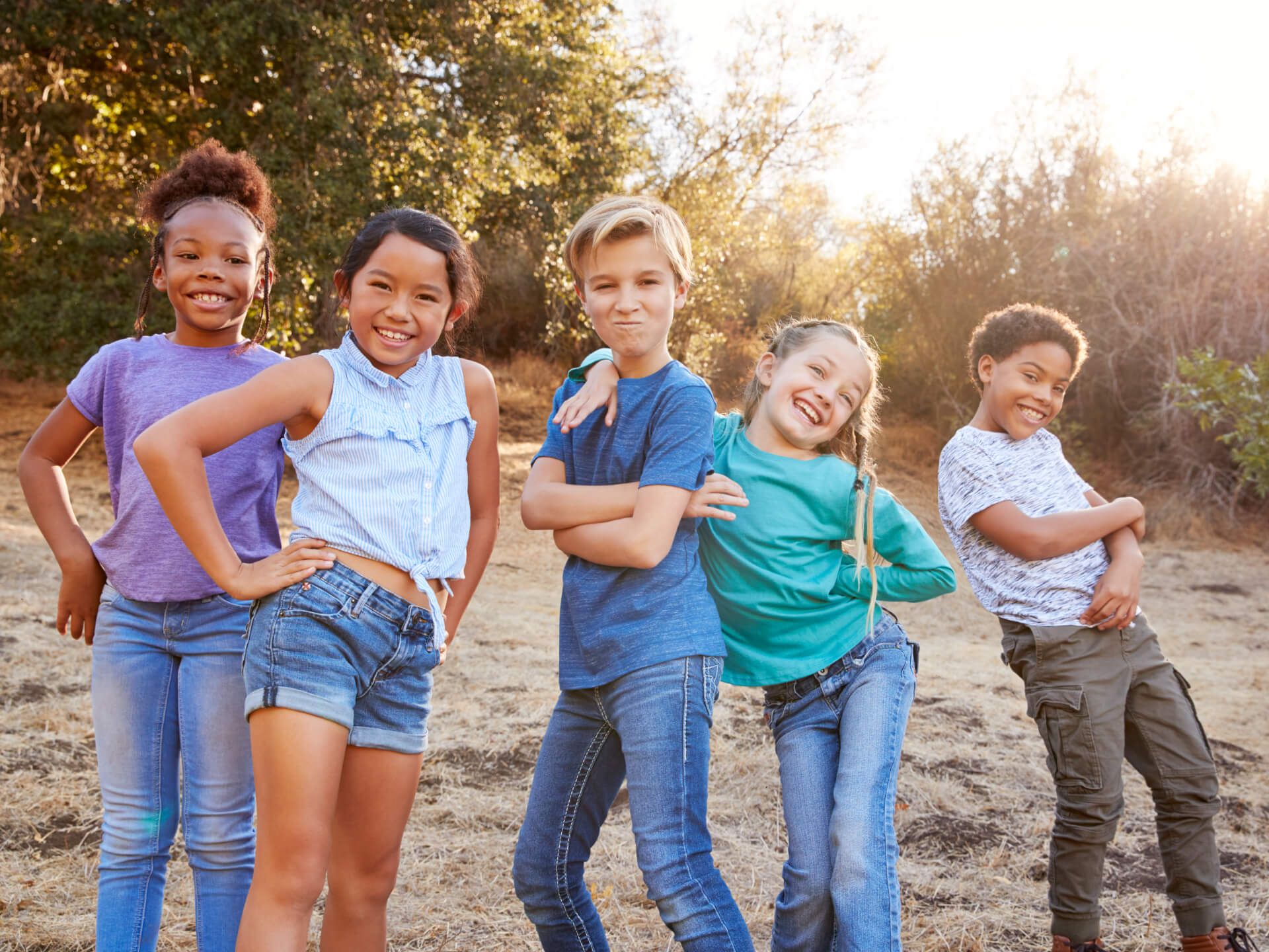 A group of five children poses playfully outdoors, standing side by side with smiles on their faces. They are dressed in casual summer clothing, including t-shirts and shorts. The background features dry grass and trees, illuminated by warm sunlight, creating a cheerful and relaxed atmosphere. The children are making fun poses, suggesting a lighthearted mood.