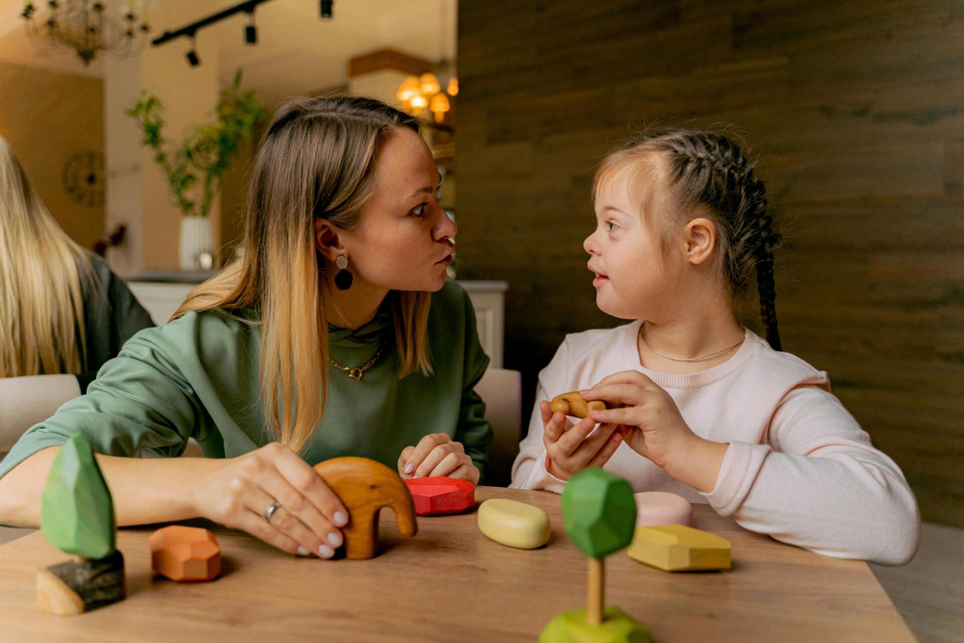 A woman and a young girl are sitting at a wooden table, engaged in a playful conversation. The woman, wearing a green hoodie and earrings, leans in towards the girl, who has hair styled in braids. The child, dressed in a light pink shirt, looks at the woman with curiosity, holding a small wooden toy. On the table, there are various colorful wooden blocks and shapes, including a green tree, a brown elephant, and other geometric forms.