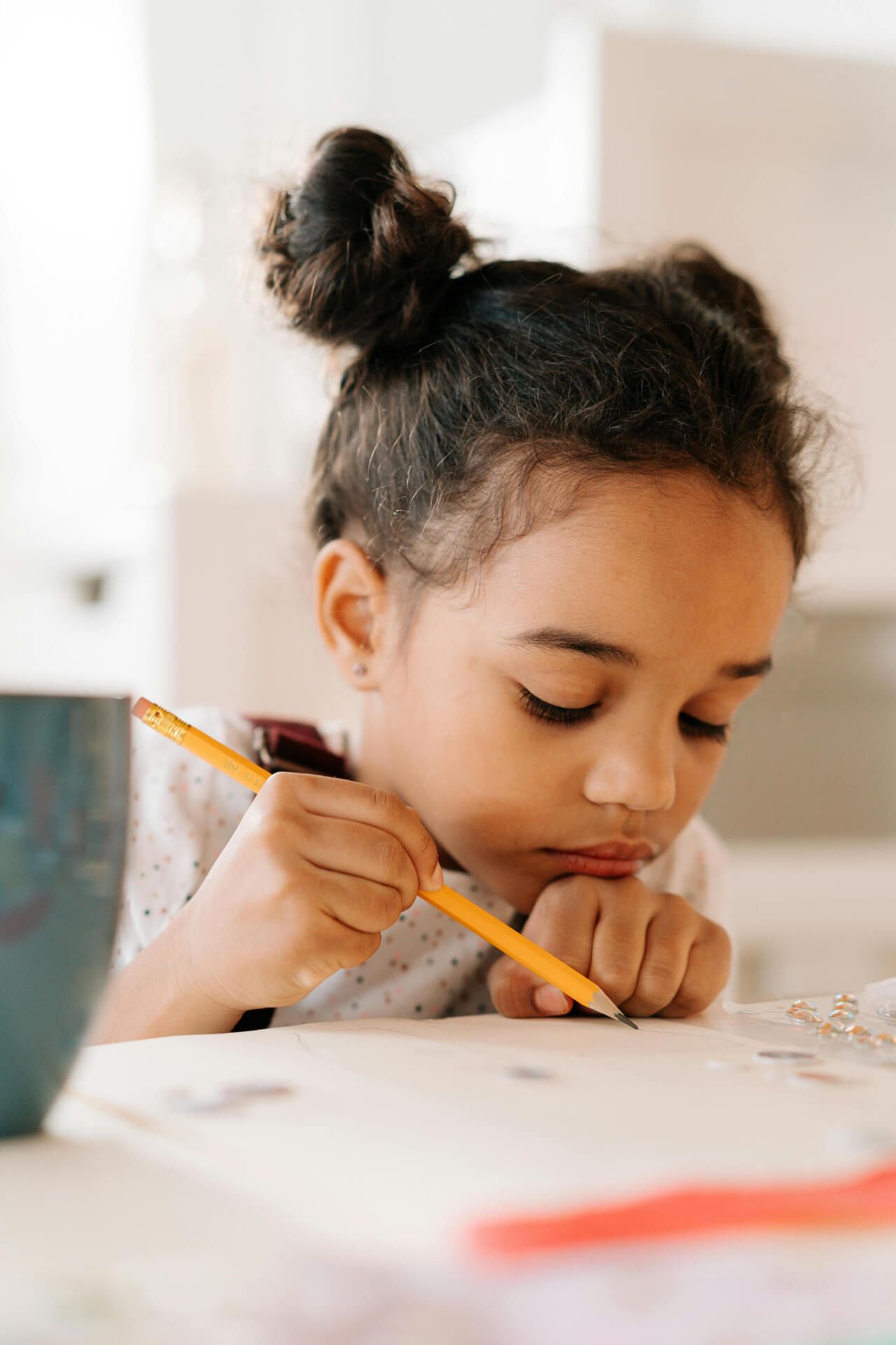 A young child with dark hair styled in two small buns is focused on drawing or crafting at a table. She holds a yellow pencil and appears to be deeply concentrated on her work, with her chin resting on her hand. The table is scattered with various crafting materials, including small shiny objects, and a colorful cup is visible in the background.